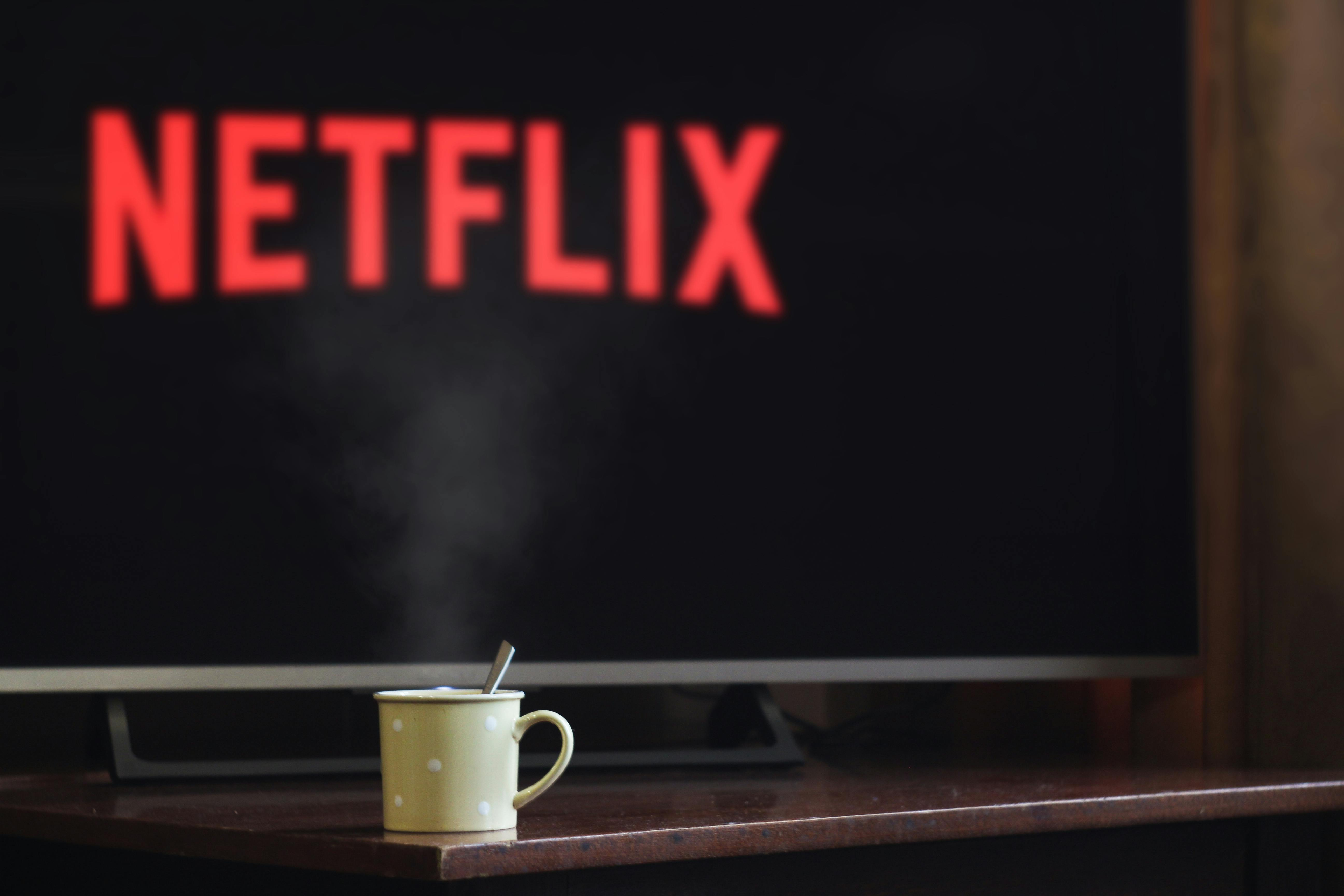 A imagem mostra uma caneca amarela com bolinhas brancas sobre uma mesa de madeira, com uma colher dentro e vapor subindo, indicando que há uma bebida quente dentro. Ao fundo, em destaque, aparece uma TV com o logotipo da Netflix em vermelho sobre um fundo preto. O foco da imagem está na caneca, com o fundo levemente desfocado, criando um clima aconchegante e intimista.